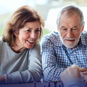 Twee oudere mensen glimlachen terwijl ze samen een schaakspel spelen in een gezellige wijkkamer van Avedan.
