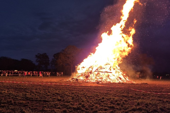 Grote vreugdevuur in een open veld bij nacht met mensen die op veilige afstand naar het vuur kijken.