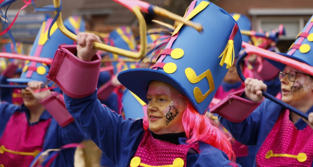 Groep mensen in kleurrijke kostuums met blauwe hoeden en muzieksymbolen tijdens een carnavalsoptocht.
