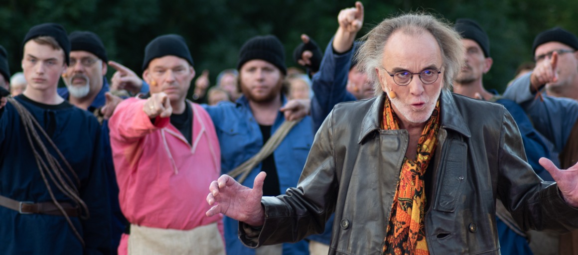 Groep mannen in donkere kleding en mutsen, één man op de voorgrond met grijze haren en bril.