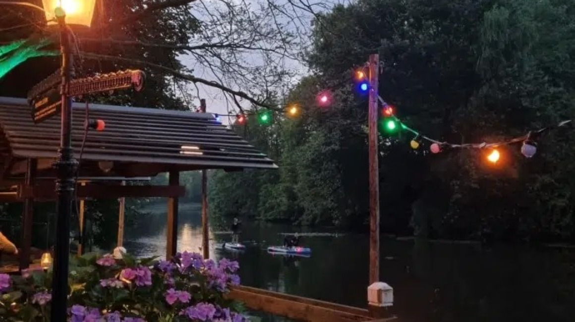 Sfeervol verlichte tuin met gekleurde lichtjes aan het water, bloemen en mensen op paddleboards bij schemering.