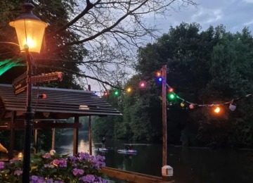 Sfeervol verlichte tuin met gekleurde lichtjes aan het water, bloemen en mensen op paddleboards bij schemering.