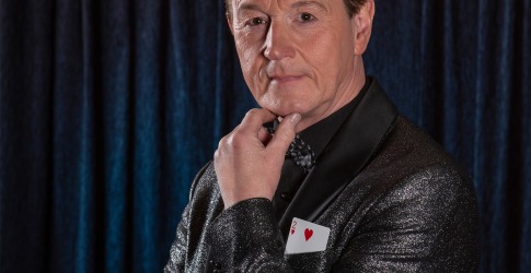 Man in een glinsterend zwart jasje met een speelkaart in de borstzak poseert voor een donkere gordijnachtergrond.