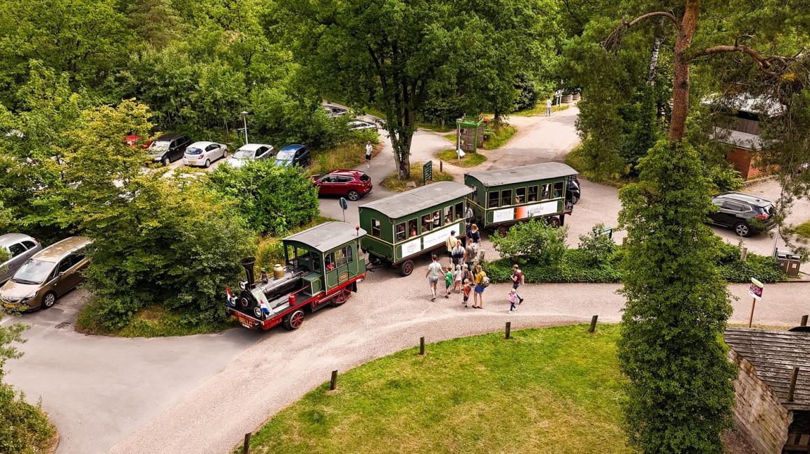 Luchtfoto van een toeristentreintje dat mensen vervoert langs een groene bosrijke omgeving met geparkeerde auto's.