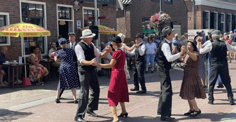 Groep mensen in vintage kleding danst op straat voor Bakkerij- & IJs-Museum op een zonnige dag.