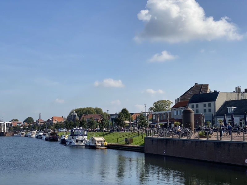 Een rustige kade met aangemeerde boten, groene grasvelden en traditionele Nederlandse huizen onder een blauwe lucht.