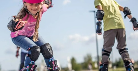 Twee kinderen dragen beschermende uitrusting en skeeleren buiten op een zonnige dag, lachend en spelend.