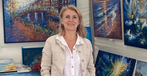 Vrouw poseert in een kunstgalerij, omringd door kleurrijke schilderijen met water- en natuurscenes.