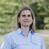 Vrouw met halflang blond haar en gouden oorbellen in een lichtblauw overhemd staat buiten in de natuur.