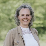 Vrouw met grijs krullend haar glimlacht en draagt een beige jasje en witte top in een groene buitenomgeving.