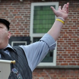 Man in ouderwetse kledij roept iets tijdens een openbaar evenement voor een bakstenen gebouw met ramen.