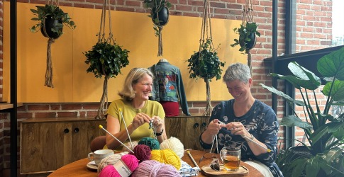 Twee vrouwen breien samen aan een tafel vol garen, planten hangen aan touwen in een gezellige ruimte.
