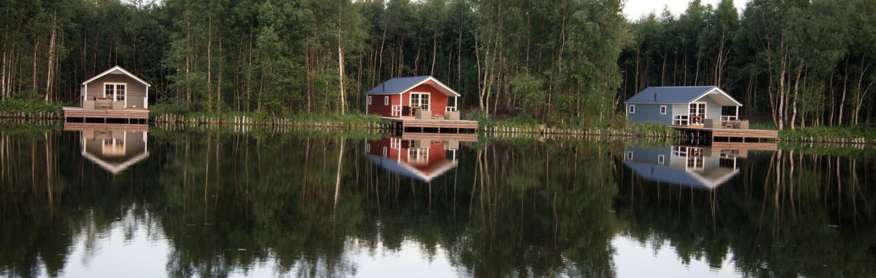 Drie huisjes aan de oever van een rustig meer met reflecties in het water en een bos op de achtergrond.
