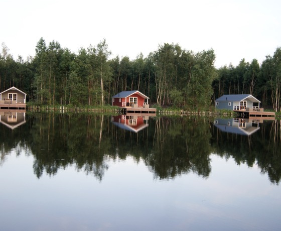 Drie huisjes aan de oever van een rustig meer met reflecties in het water en een bos op de achtergrond.