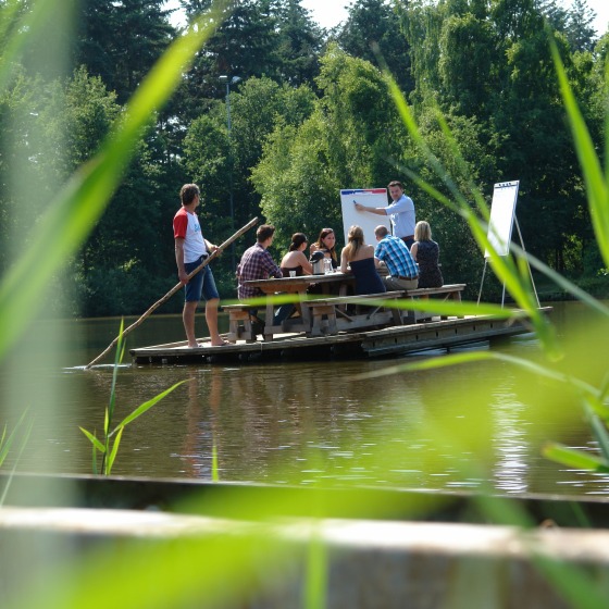 Een groep mensen houdt een vergadering op een vlot op het water, omgeven door groene bomen en gras.
